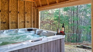 A hot tub with a wooden exterior, surrounded by a wooden deck, and a view of a forest through a large window. - Discounts 3+N 4BR Cabin Sleeps 10 Hot Tub, Game Room, Fire Pit, Fenced Yard-Pets (Blue Ridge)