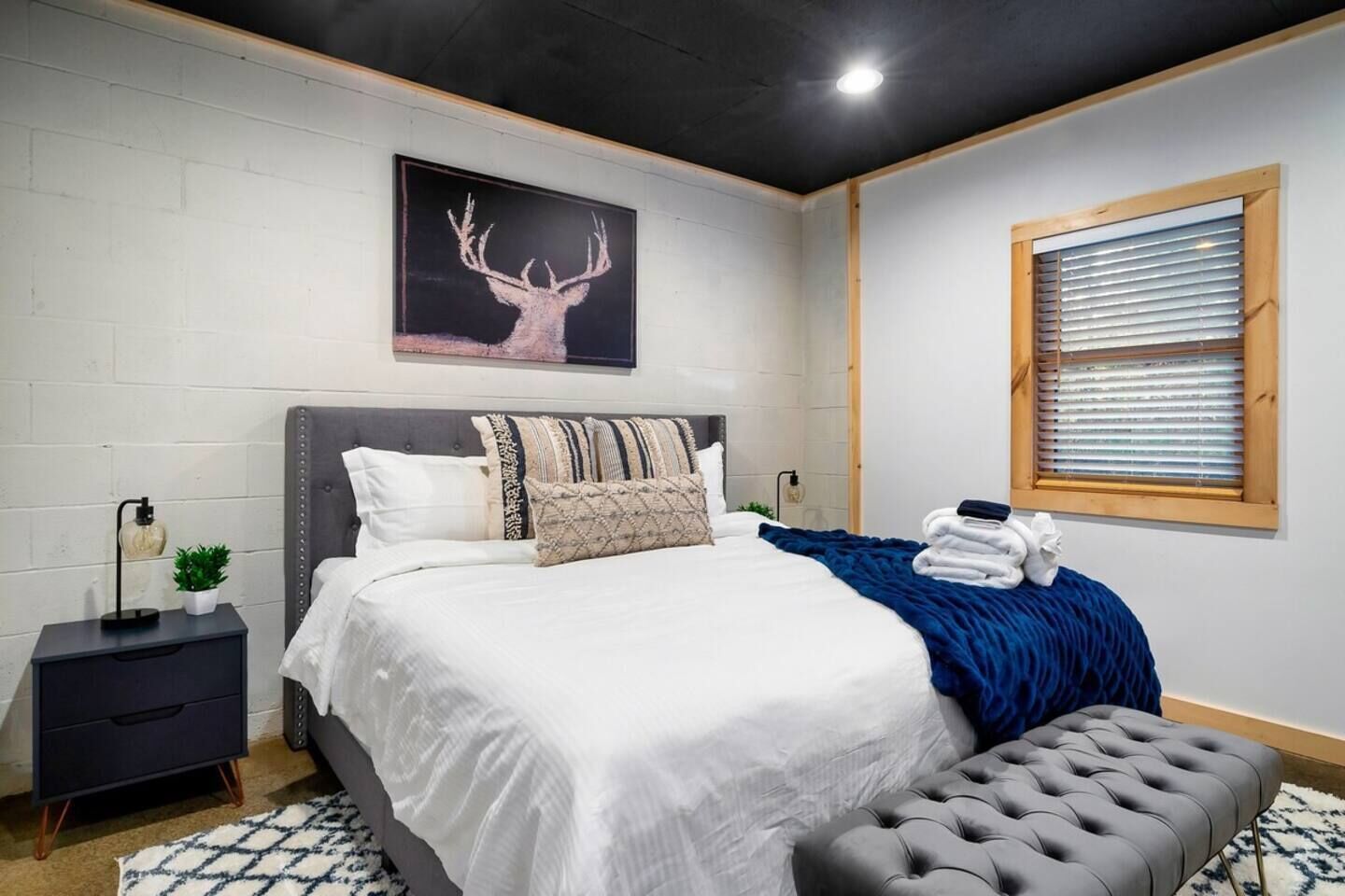 A modern bedroom with a large bed, a nightstand, a window with blinds, and a wall-mounted picture of a deer.