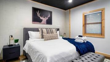 A modern bedroom with a large bed, a nightstand, a window with blinds, and a wall-mounted picture of a deer.