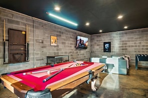 Une table de billard avec des billes et des queues, un téléviseur fixé au mur, et un coin salon avec un canapé et des fauteuils.