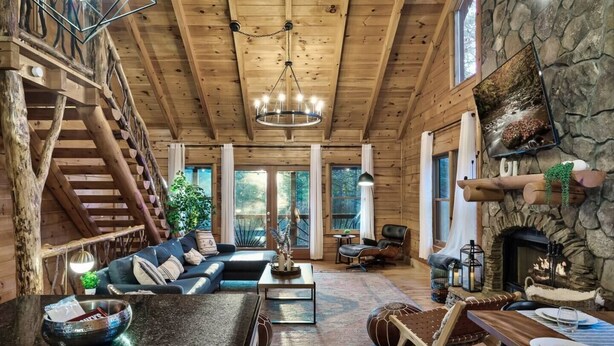 A spacious log cabin interior with a large living area, a stone fireplace, a dining area, and a staircase leading to an upper level. - Discounts 3+N 4BR Cabin Sleeps 10 Hot Tub, Game Room, Fire Pit, Fenced Yard-Pets (Blue Ridge)