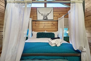 Une chambre à coucher avec un lit, des rideaux blancs, un tableau encadré représentant un taureau et une vue sur l’extérieur.