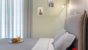 A modern hotel room with a bed, a bedside table with a book and a flower, a wall-mounted air conditioner, and a window with curtains. - Suite 06 - Smart Cozy Suites - Cozy, elegant and near the metro (Kallithea, Athens)