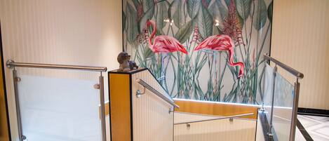 Vestíbulo de hotel con escalera, barandilla de vidrio y un mural que muestra flamencos.