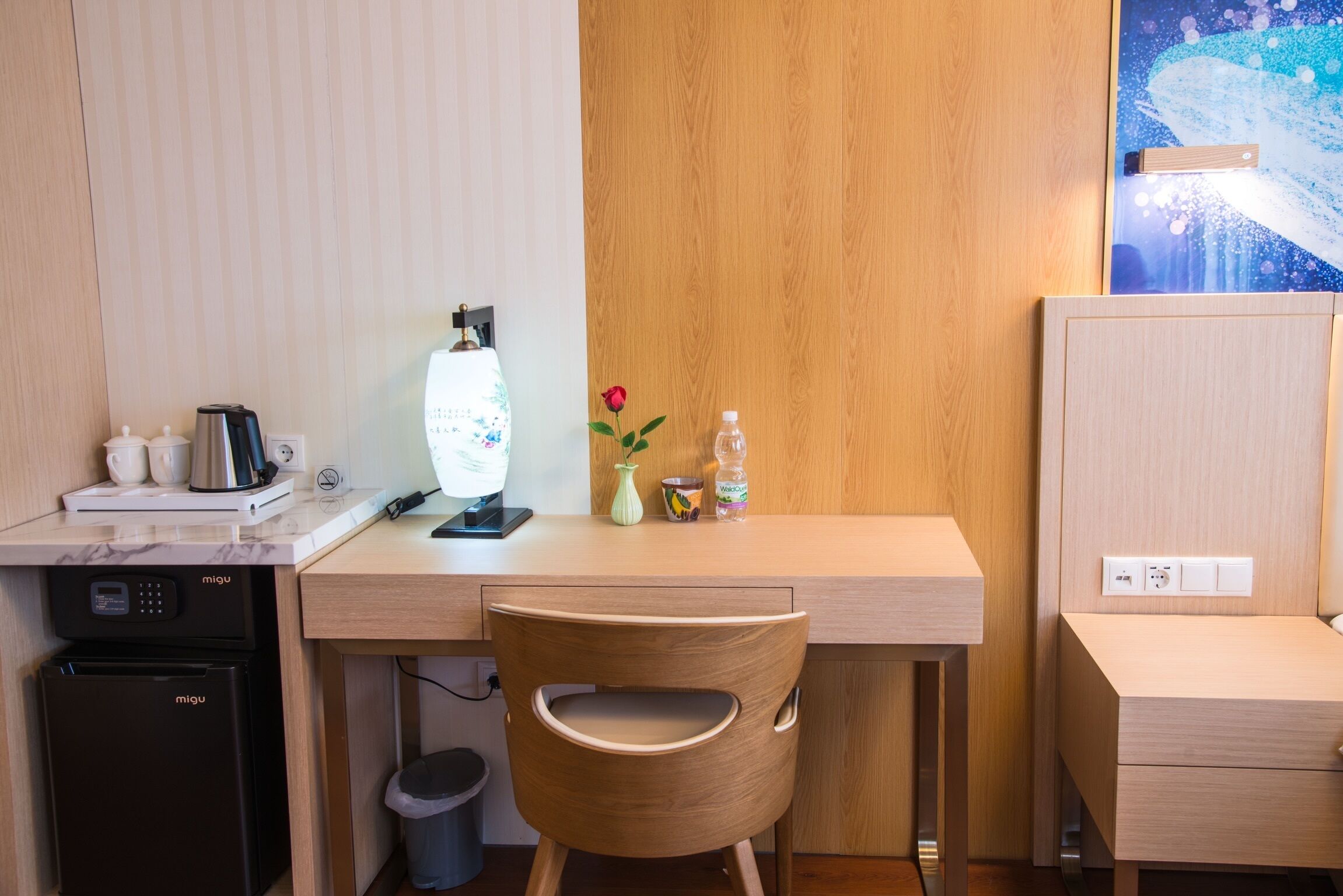 A compact hotel room with a wooden desk, a small refrigerator, a microwave, a trash bin, and a single chair.