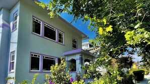 Uma casa de dois andares com fachada azul clara e detalhes em roxo, cercada por vegetação exuberante e uma cerca de metal preto.