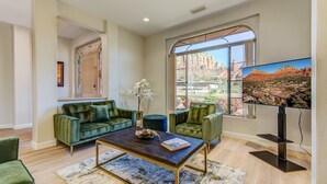 A spacious living room with a large window offering a view of red rock formations, a comfortable green sofa set, a coffee table, and a flat-screen TV. - Experience luxury at Sedona Chapel Area Charm, a spacious 4BR home with stunning red rock views. (Sedona)