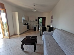 A living room with a sofa, a coffee table, and a dining area with chairs and a table. There is a kitchen area with cabinets and a sink.
