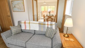A grey sofa with patterned cushions, a wooden side table with a lamp, and a dining area with a brick wall and wooden chairs. - Larch Lodge (Totland Bay)