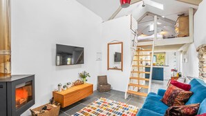 A modern living room with a fireplace, a blue sofa, a colorful rug, and a loft bedroom above. - Luxury self-contained cottage for two with wood fired hot tub (Staverton)
