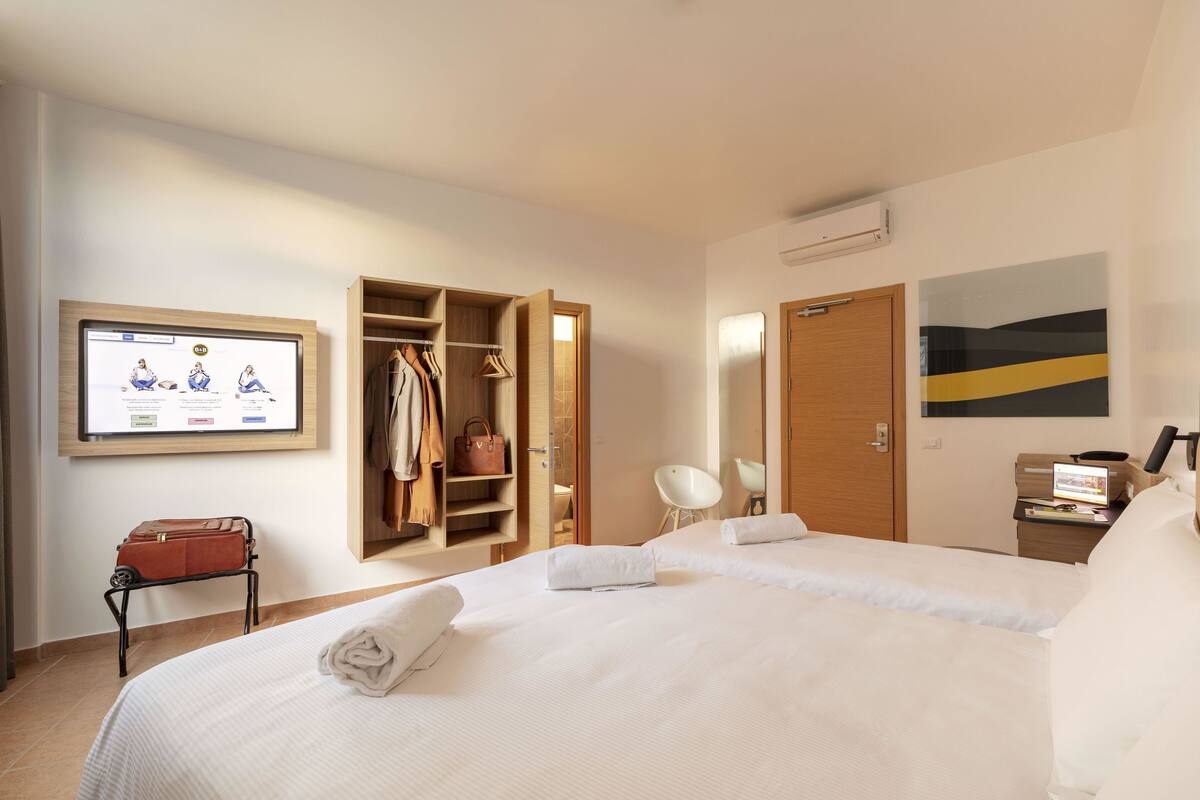 Quarto de hotel com uma cama grande, um guarda-roupa, uma escrivaninha com um computador e uma televisão na parede.