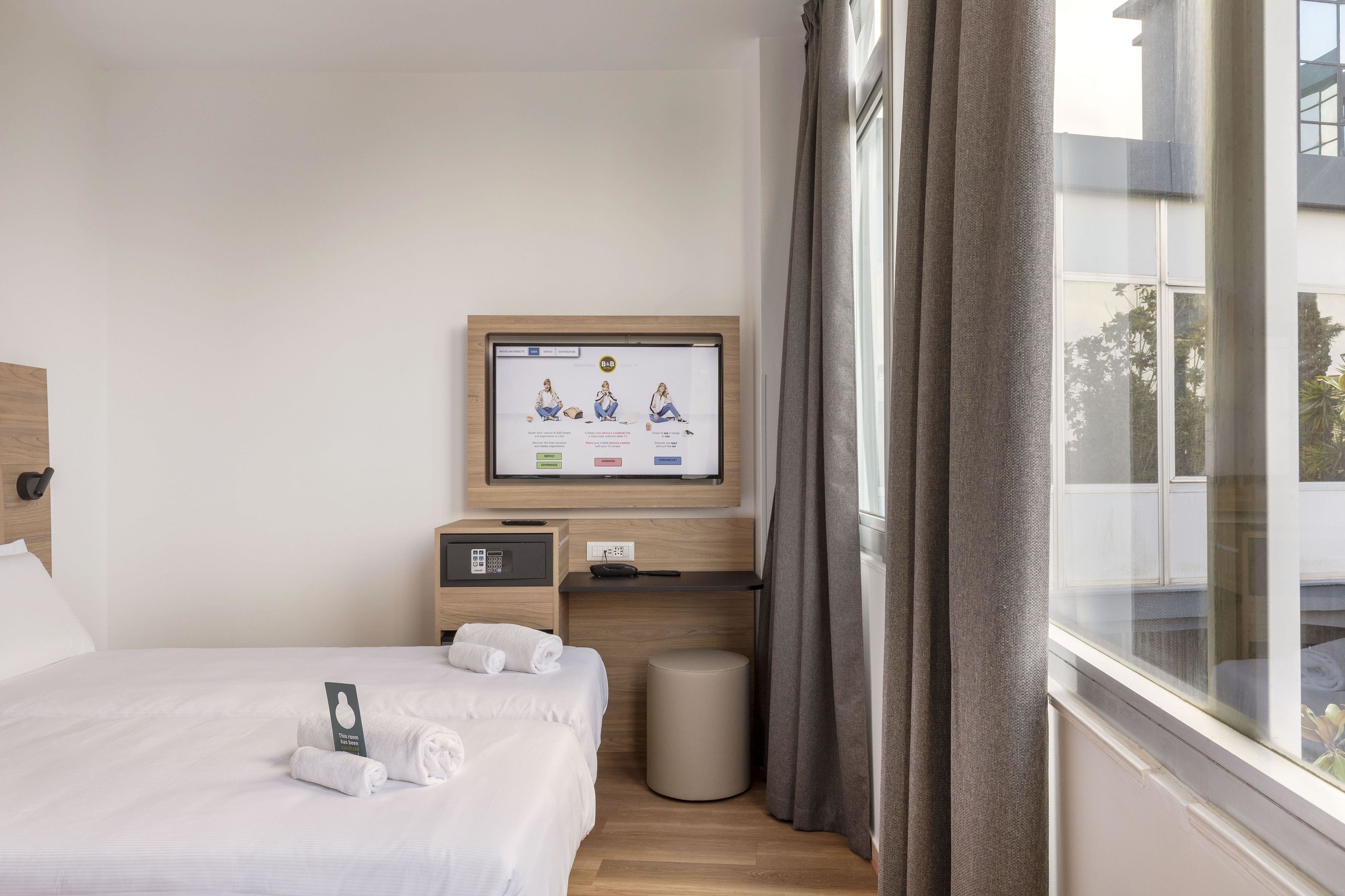 A hotel room with a bed, a TV mounted on the wall, a small desk, a trash bin, and a window with curtains.