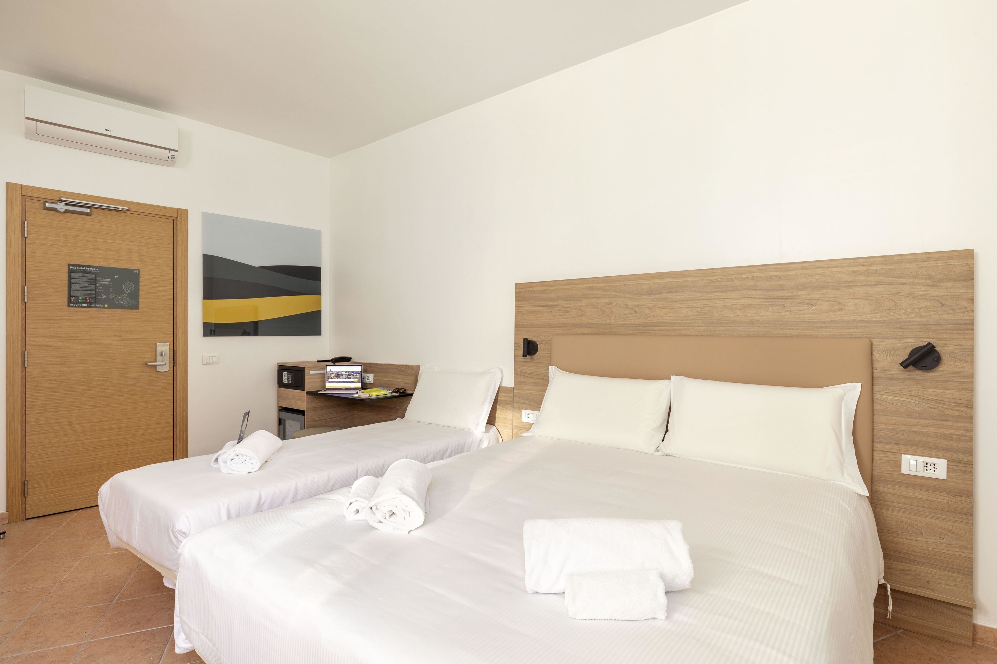 A hotel room with two beds, a wooden headboard, a desk with a computer, and a painting on the wall.