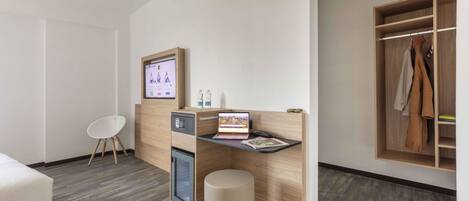 A hotel room with a wooden desk, a laptop, a small refrigerator, a white chair, a round ottoman, and a wardrobe.