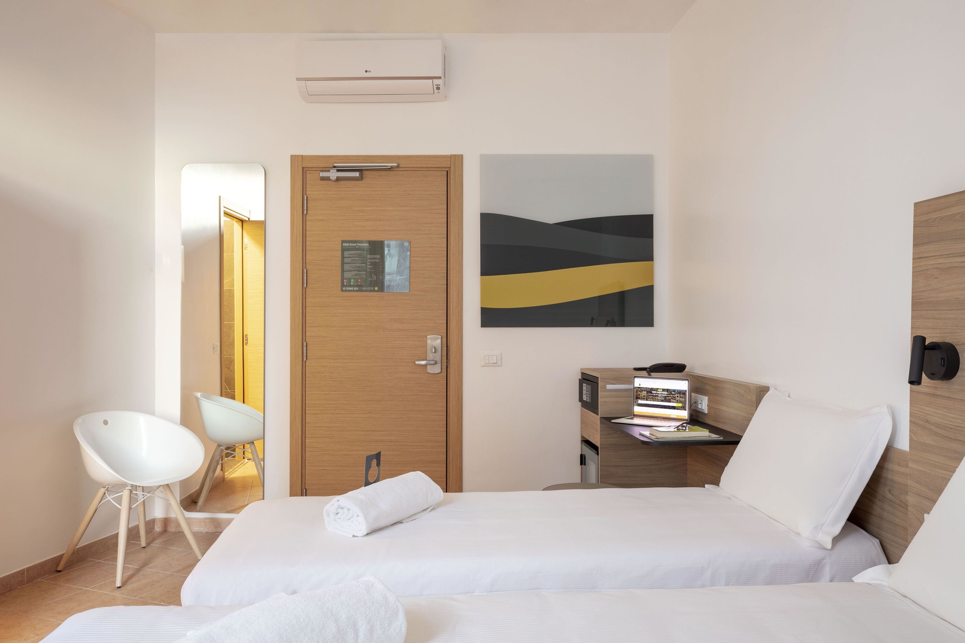 A hotel room with two beds, a desk with a computer, a chair, and a wall-mounted air conditioner.