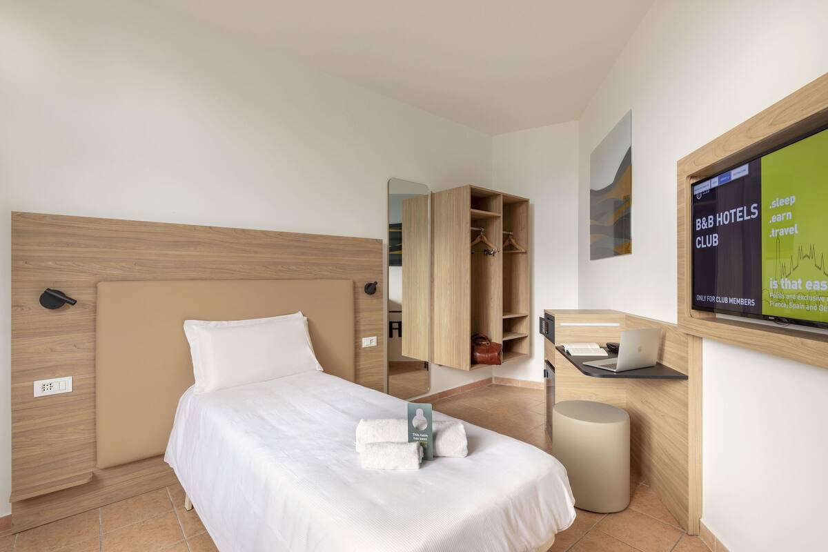 Quarto de hotel com cama, secretária com laptop, espelho e guarda-roupa.
