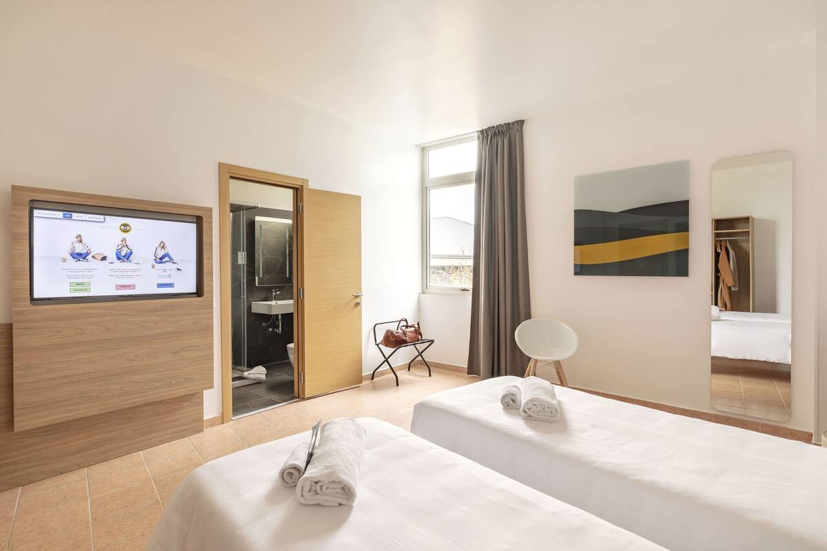 Quarto de hotel com duas camas, televisão, espelho e janela com cortinas.