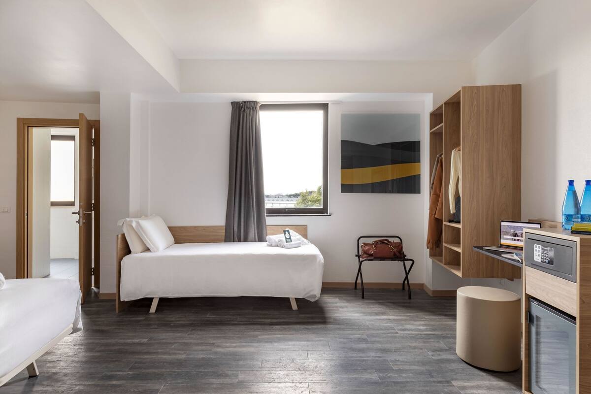 Quarto de hotel moderno com cama, escrivaninha, cadeira, mesinha, lixeira e janela com vista.