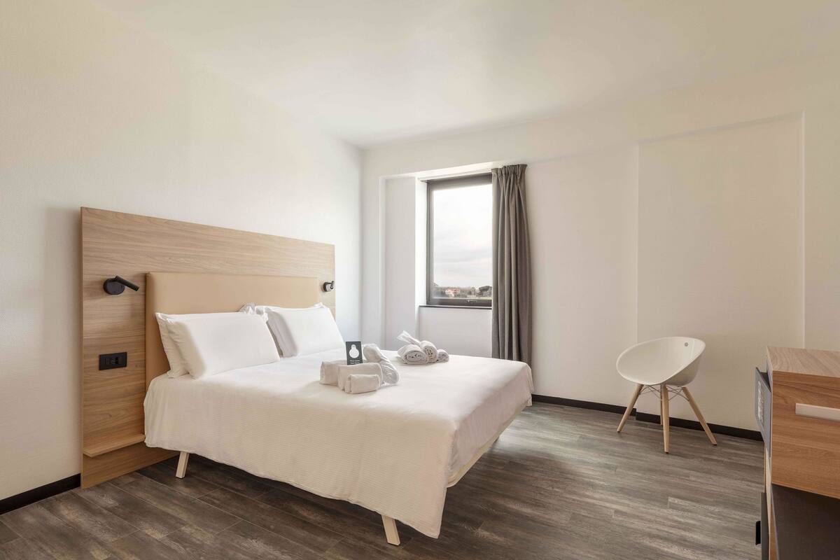 Quarto de hotel com cabeceira de madeira, cama com roupa de cama branca, uma pequena escrivaninha, uma cadeira e uma janela com cortinas.
