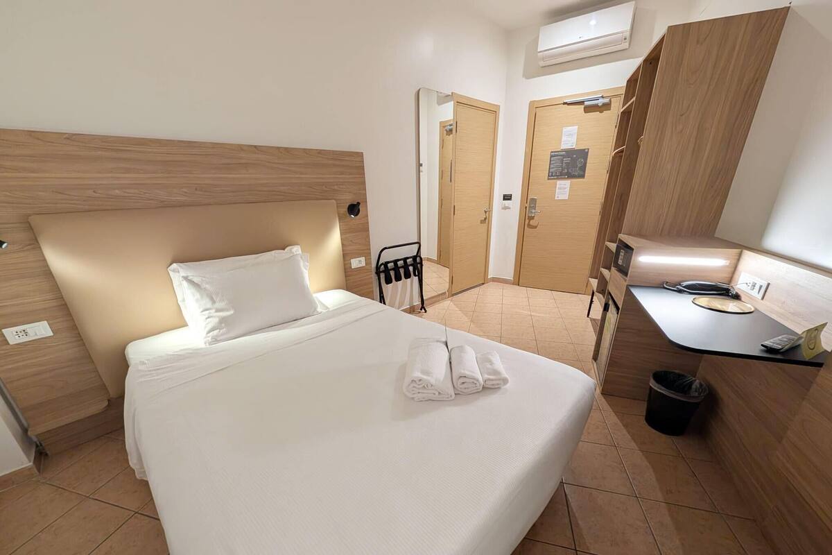 Quarto de hotel com uma cama, uma escrivaninha, uma cadeira e um cesto de lixo.