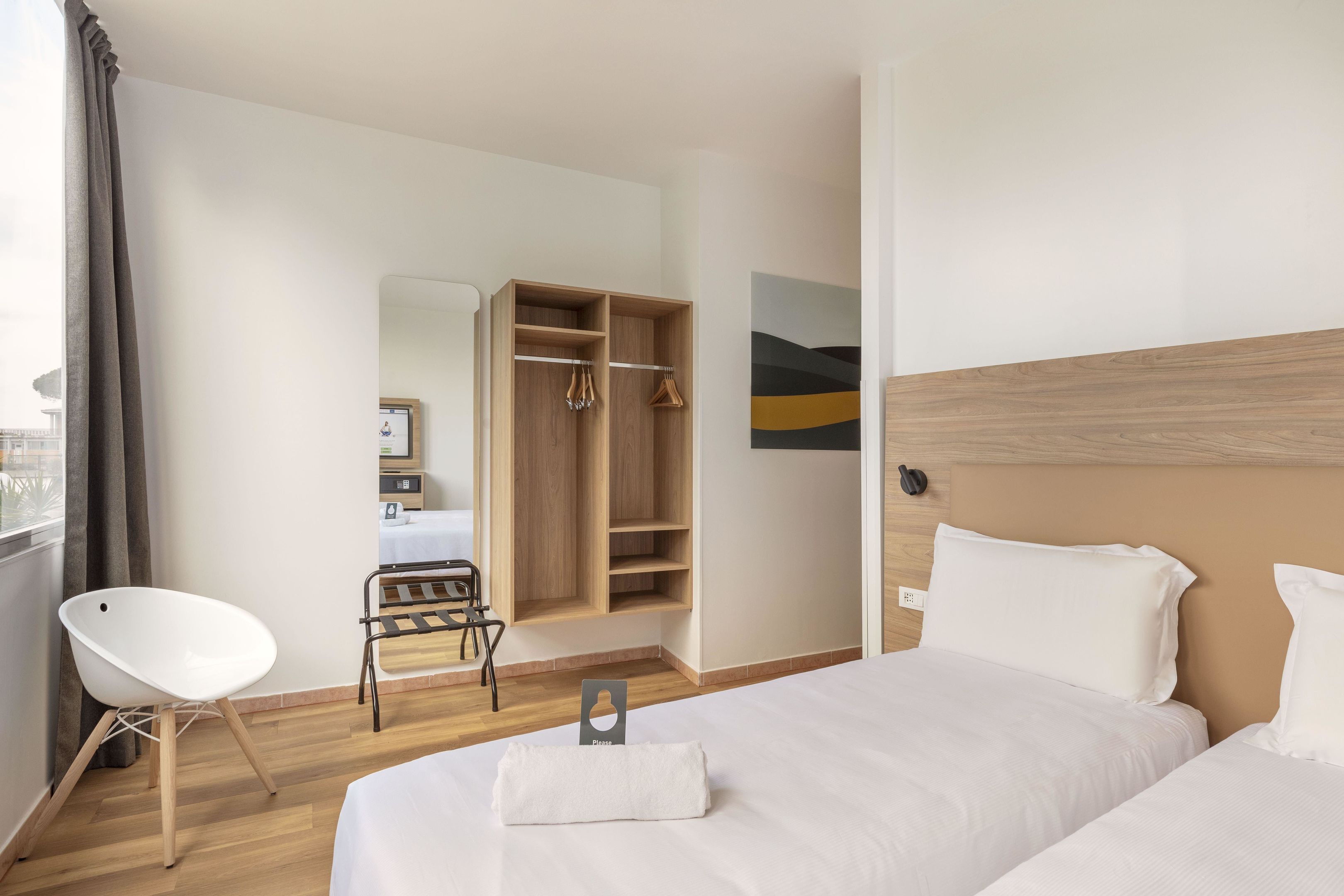 A hotel room with two beds, a wooden wardrobe, a chair, and a view of the city.