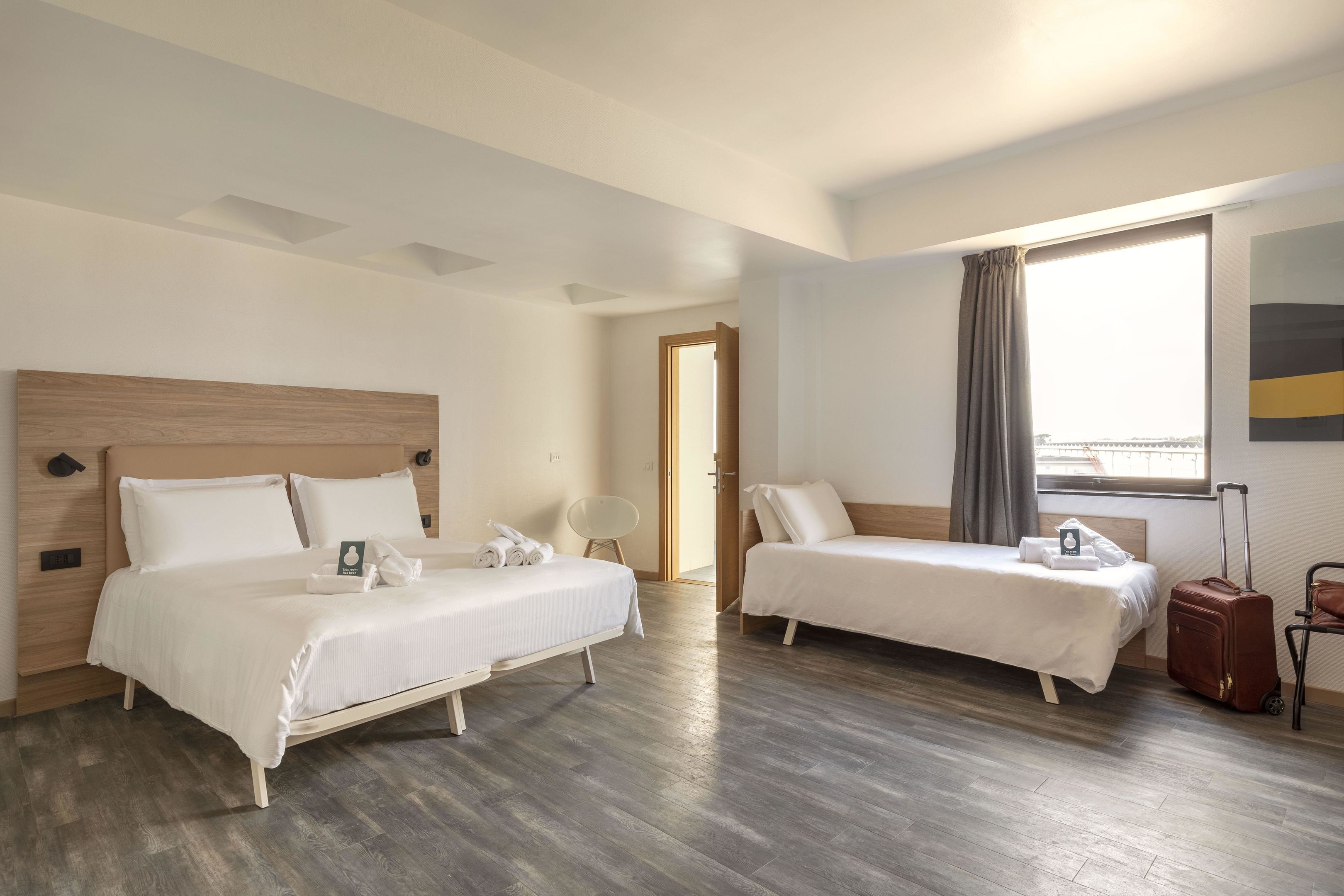 A hotel room with two beds, a wooden headboard, a large window with curtains, and a suitcase.