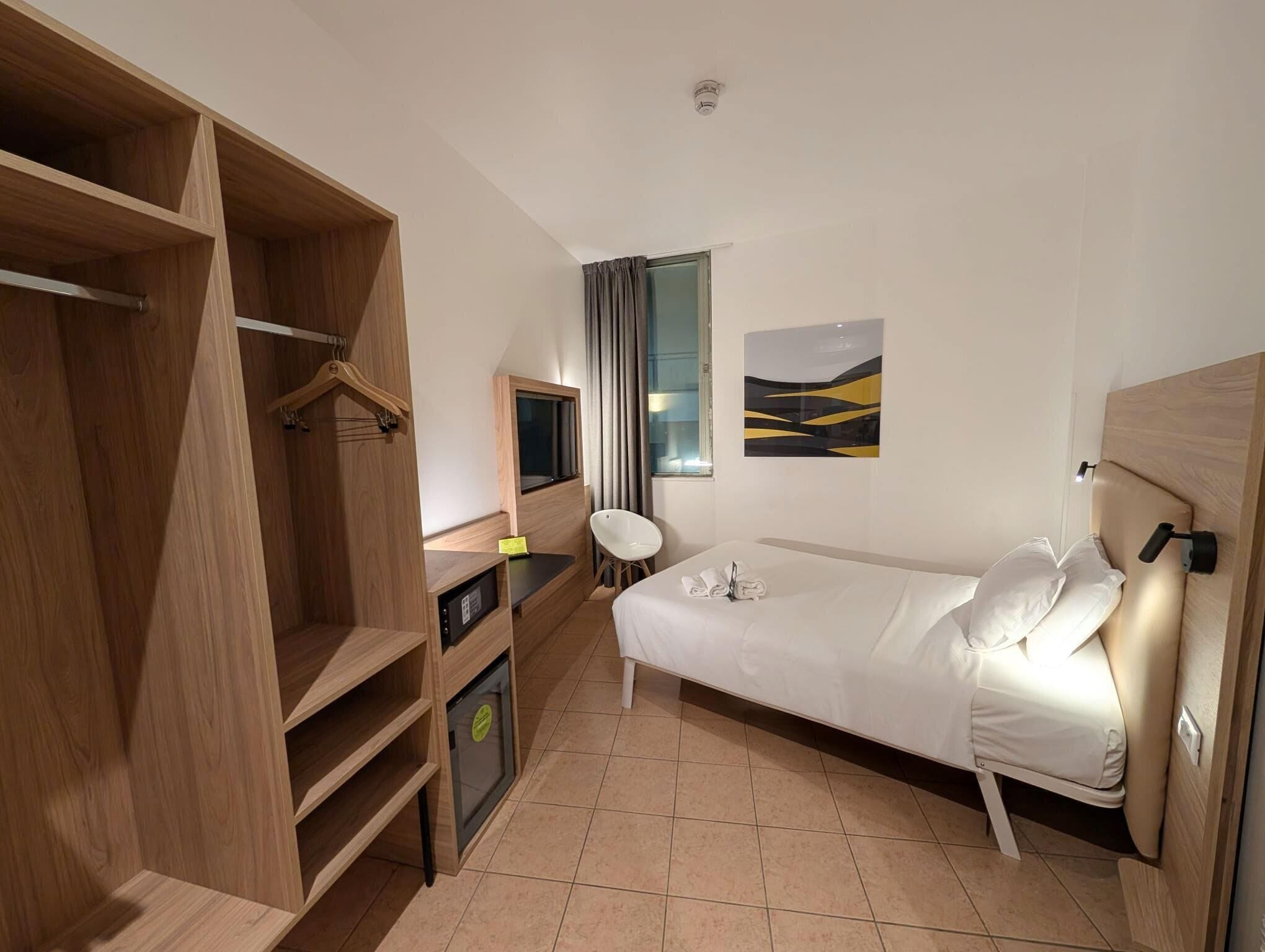 Habitación de hotel con cama, escritorio con computadora, armario y ventana con cortinas.