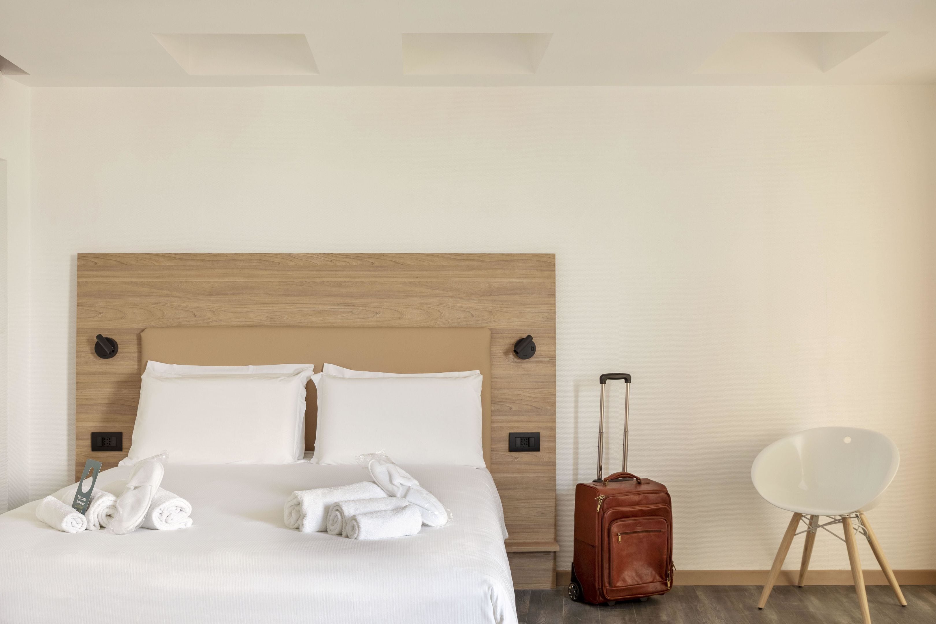 A hotel room with a wooden headboard, a neatly made bed with white linens, a brown suitcase, and a white chair.