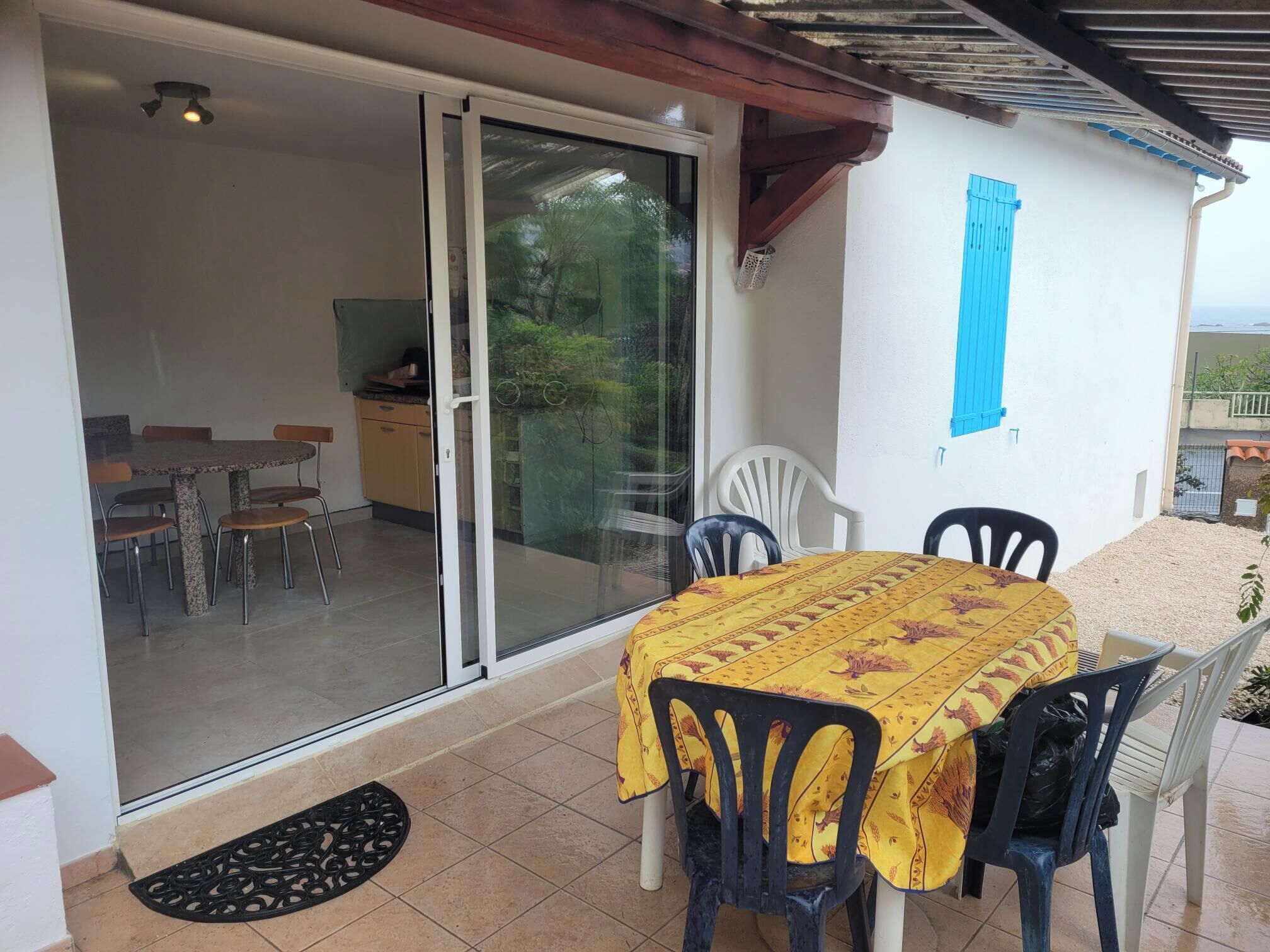 Een terras met een tafel en stoelen, een schuifpui naar een keuken, en een blauw luik aan de muur.