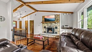A spacious living room with a stone fireplace, leather sofas, wooden beams, and a view of the outdoors. - Outdoor Pool ● Near Boone & Elk ● Fire Pit ● Games ● Kids Bunks (Banner Elk)