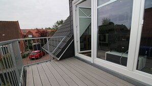 A wooden deck with a metal railing, a red car parked in the background, and a building with large windows and a sloped roof.