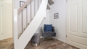 A staircase with a white handrail and wooden steps leading to a landing. A grey armchair with a blue cushion is placed on the landing, next to a small grey ottoman. The floor is covered with wooden parquet.