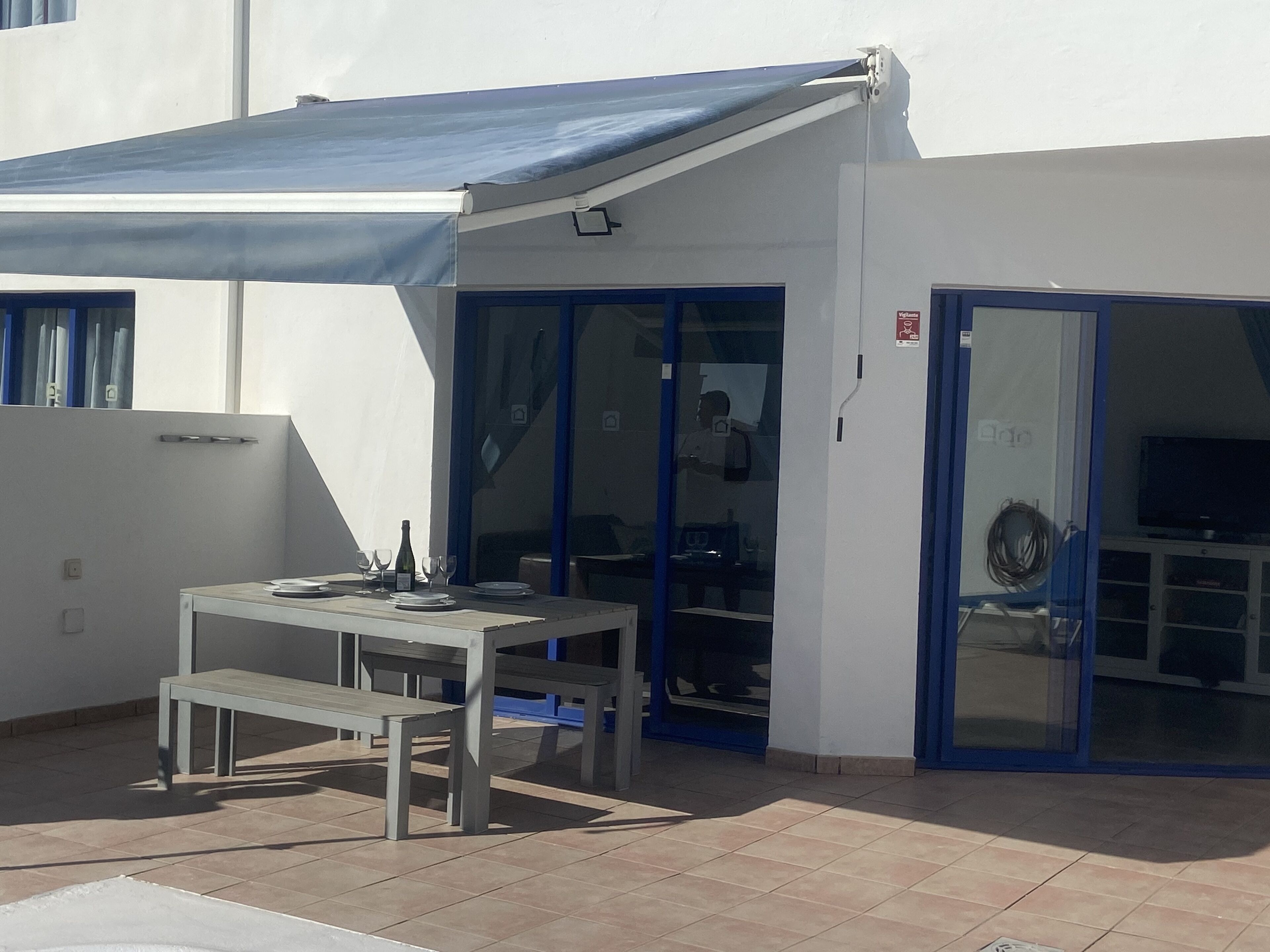 En udendørs terrasse med et bord og stole, et blåt markise og en skydedør, der fører til et indendørs rum med et fjernsyn.