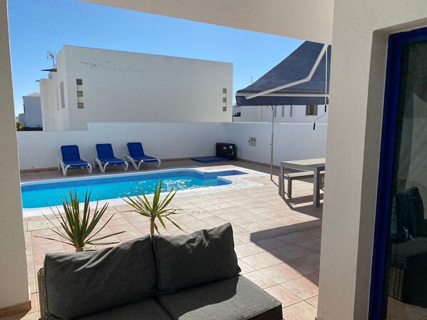 Un área de piscina con sillas reclinables azules, un sofá gris y suelo de baldosas.