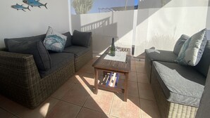 Zona d'estar a l'aire lliure amb llum solar, mobles de vímet, una taula petita amb una ampolla i vasos, i peixos decoratius a la paret.