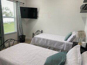 Una habitación con dos camas individuales, un televisor fijado a la pared, una ventana con cortinas y una mesita de noche con lámpara.