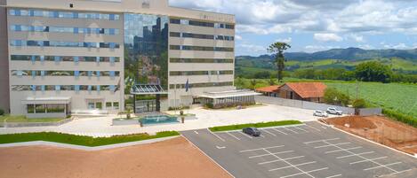 A modern hotel with a large parking lot, a swimming pool, and a view of green hills in the background.