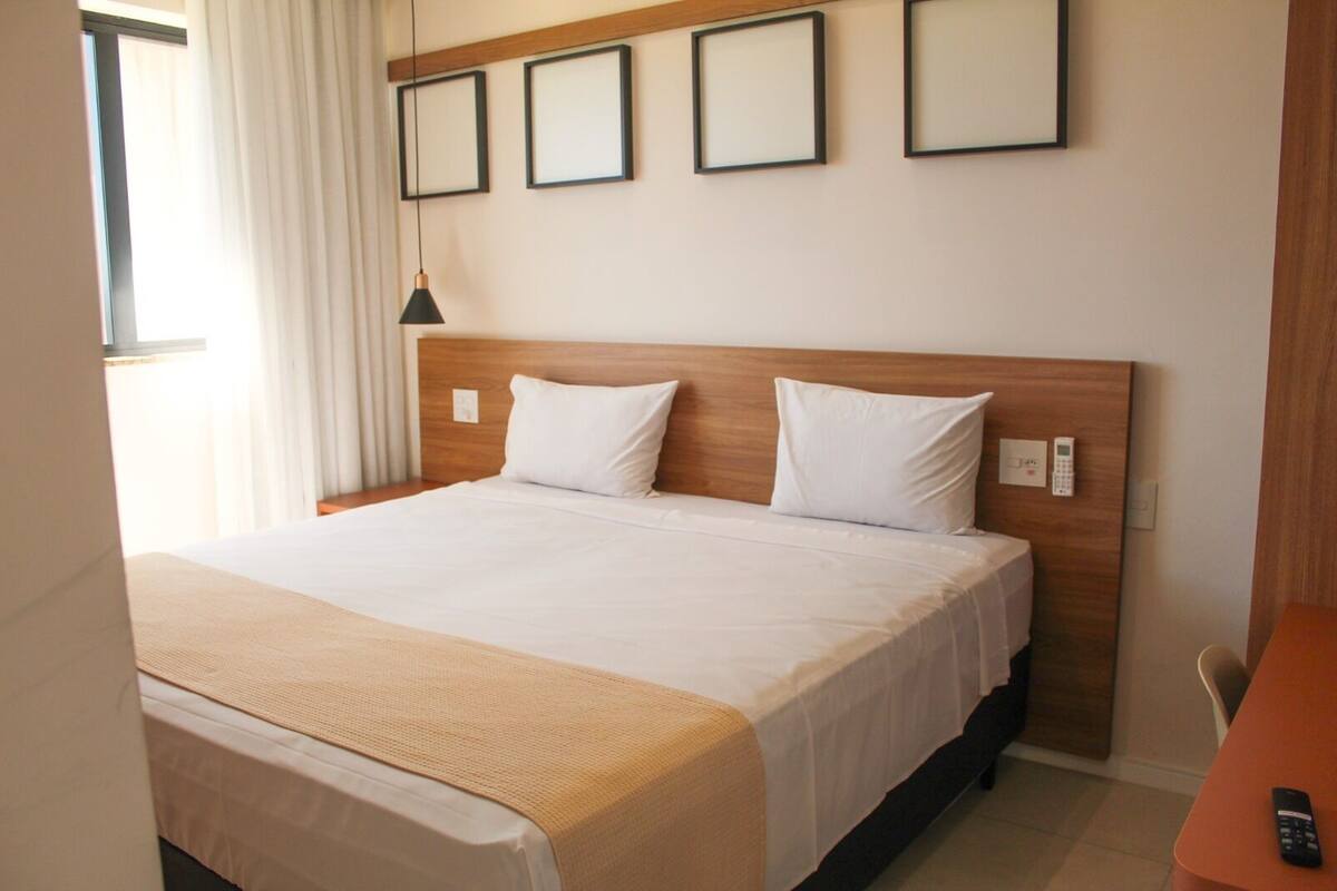 A hotel room with a large bed, a wooden headboard, a bedside table, a remote control, and a wall-mounted thermostat.
