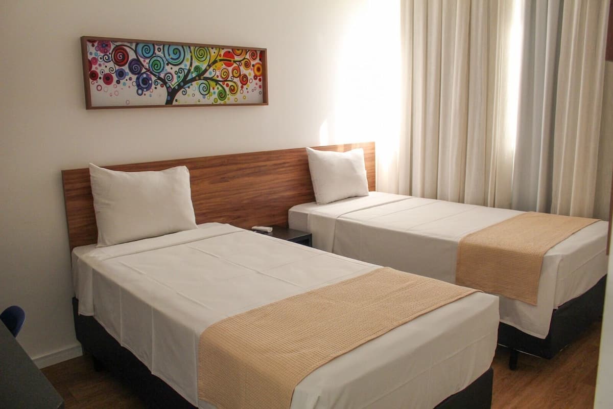 A hotel room with two beds, a wooden headboard, a colorful abstract painting, and a window with curtains.