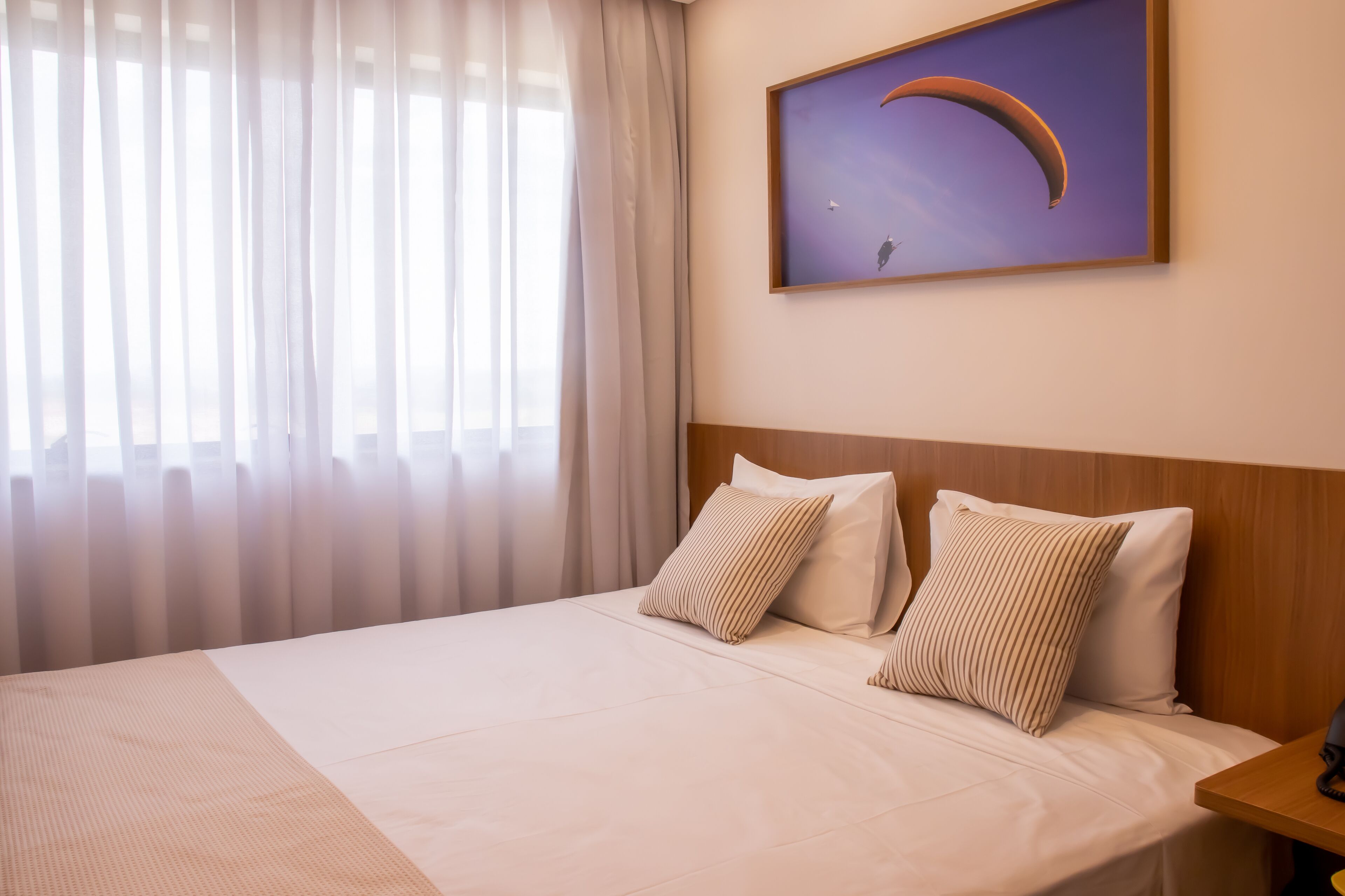 A hotel room with a bed, two pillows, a wooden headboard, a nightstand, a framed picture on the wall, and sheer curtains on the window.