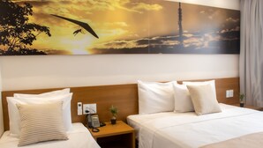 Hotelkamer met twee bedden, een houten hoofdbord, een nachtkastje met een telefoon en een plant, en een groot schilderij van een zonsondergang met een vogel en een toren.