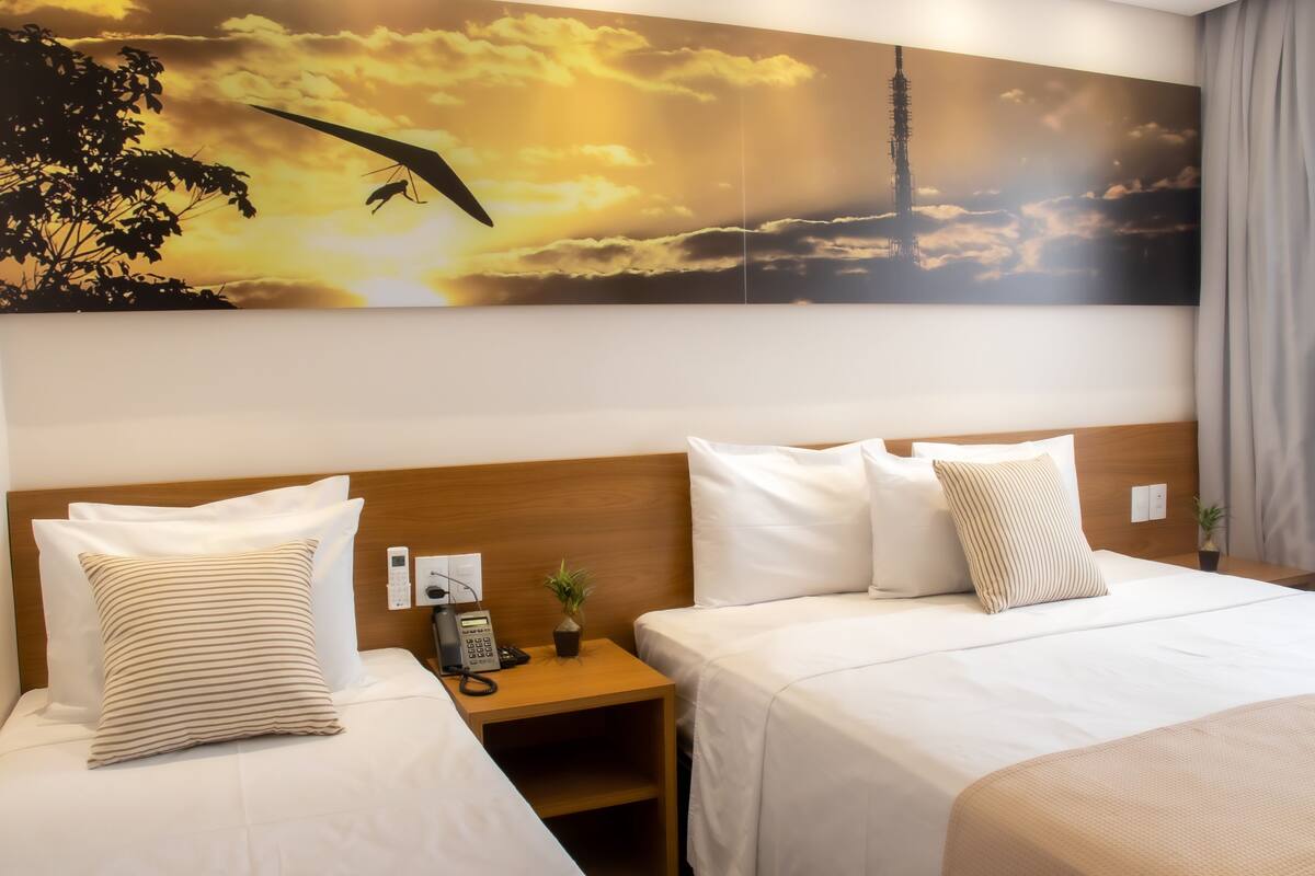 A hotel room with two beds, a wooden headboard, a nightstand with a phone and a plant, and a large painting of a sunset with a bird and a tower.