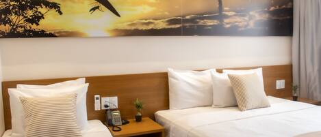 A hotel room with two beds, a wooden headboard, a nightstand with a phone and a plant, and a large painting of a sunset with a bird and a tower.