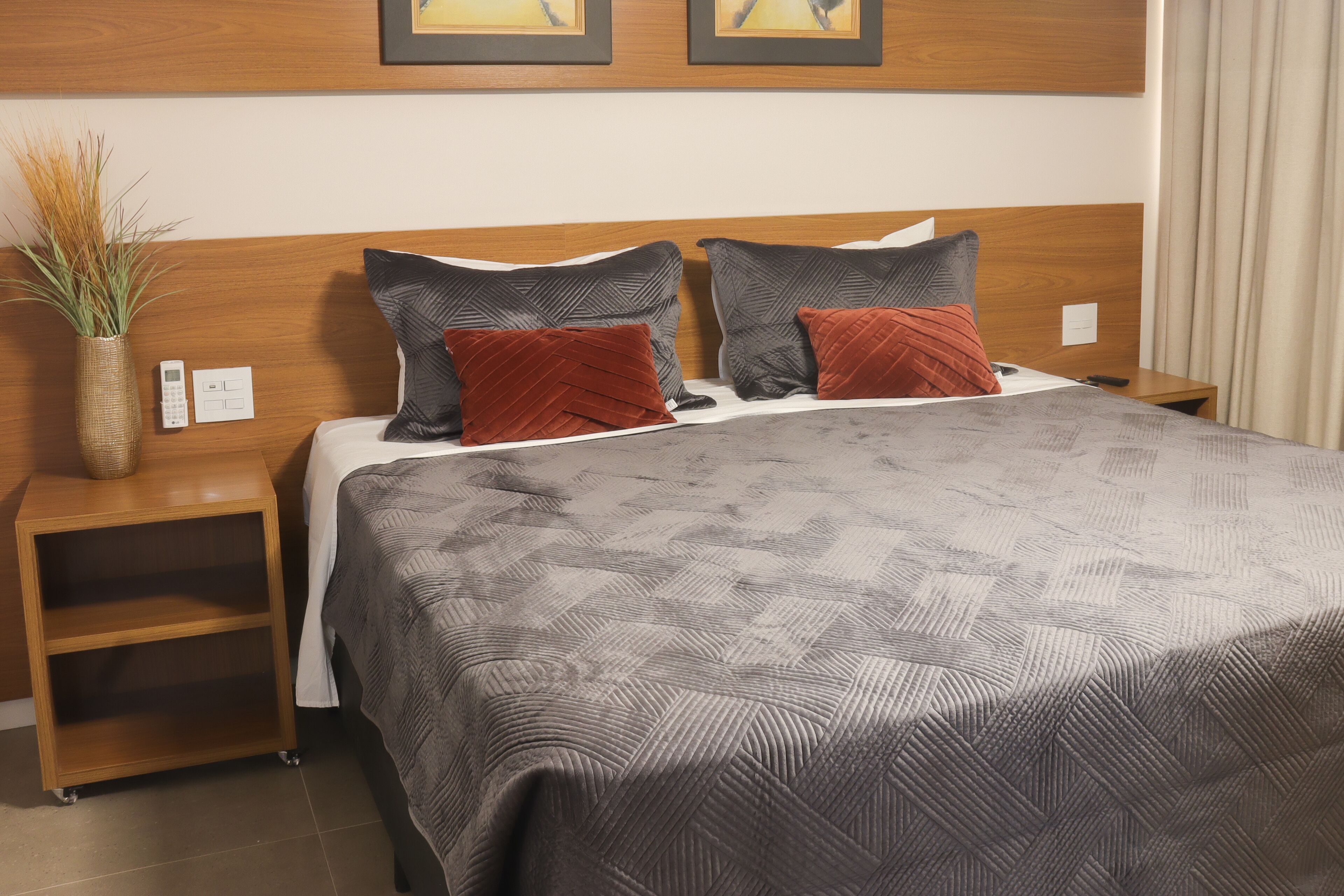 A neatly made bed with a patterned bedspread, two pillows, a bedside table with a vase of plants, and a wall-mounted thermostat.