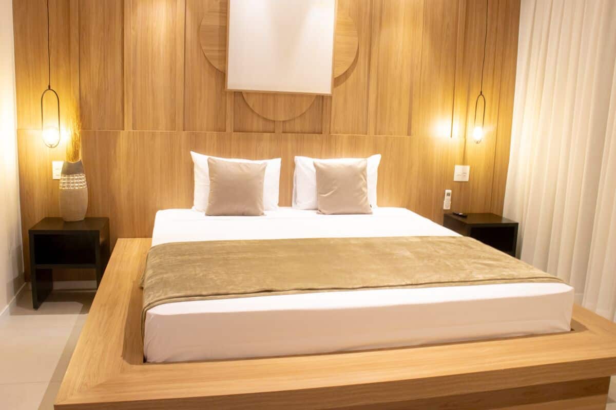 A modern hotel room with a wooden headboard, two bedside tables, a large bed with white linens, and wall-mounted lighting.