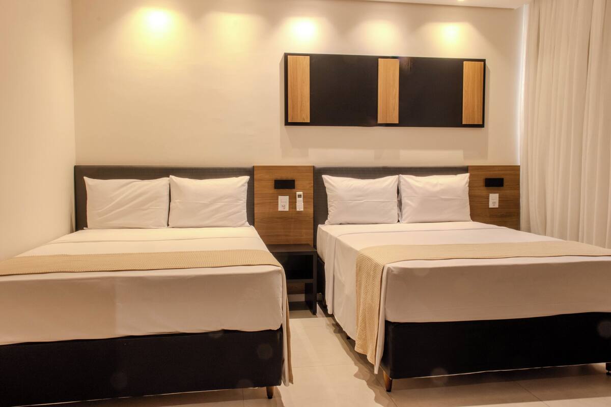 A hotel room with two single beds, a wooden headboard, a flat-screen TV mounted on the wall, and a bedside table with a lamp.