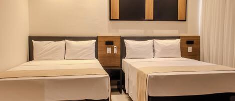 A hotel room with two single beds, a wooden headboard, a flat-screen TV mounted on the wall, and a bedside table with a lamp.