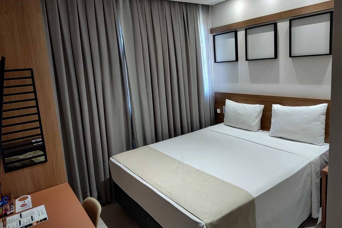 A hotel room with a large bed, grey curtains, a wooden headboard, and a small table with a chair.