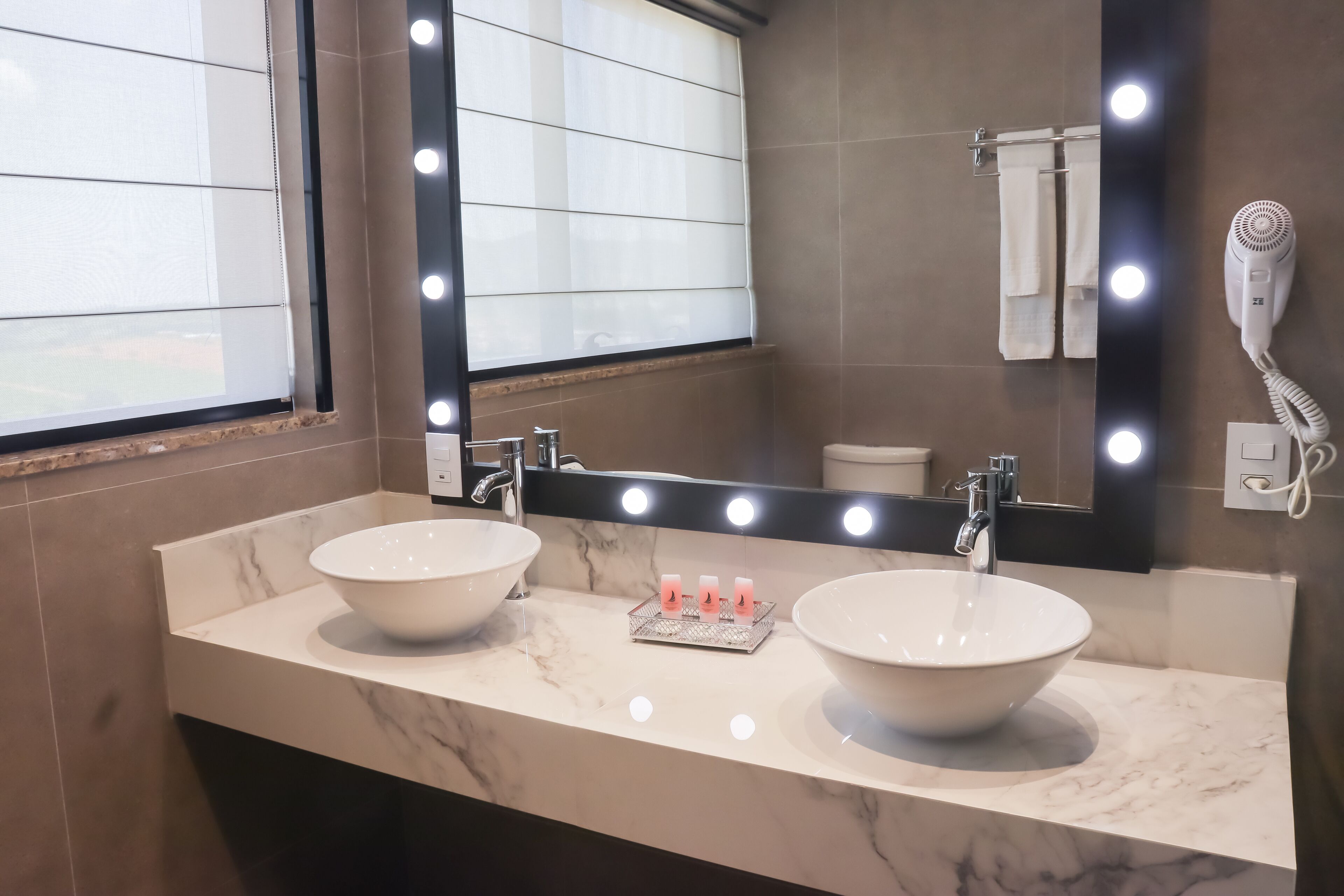 A bathroom with two sinks, a mirror with lights, a hairdryer, and a towel rack.