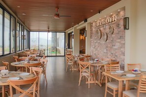 A spacious dining area with wooden tables and chairs, large windows offering a view of palm trees and a distant landscape, and a brick wall with the text "Terminal 22". - Porto das Asas Park Hotel (Andradas)